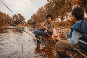 Happy little family enjoying snack while fishing. Sitting in the river on fishing chairs with fishing rods in water